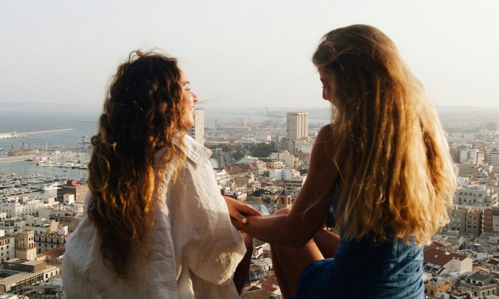 Two young girls, sitting facing each other and talking with a city in the background in front of them.
